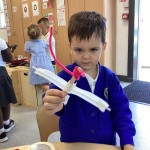 A young boy, wearing a blue cardigan over a white collared shirt, is holding up a handmade toy airplane with white wings and a pink propeller-like attachment. He is looking at the camera.