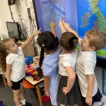 Four young children, also in white polo shirts, are standing in front of a large interactive screen, all pointing at different parts of a map that appears to show the UK and surrounding areas. This is likely in a classroom.