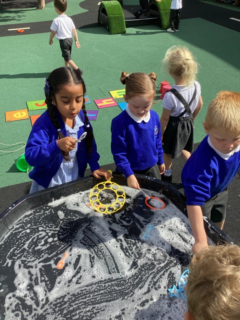 Several children are gathered around a large black tub filled with soapy water and bubbles. Two girls, both wearing blue tops, are actively playing with bubble wands in the water. Another child's arm is visible on the right, also engaging with the water.