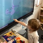 A young child with light brown hair, wearing a white polo shirt, is pointing at a large interactive screen displaying what looks like a map or geographic information. There are various colorful objects on the table in front of the screen. This appears to be in a classroom setting.