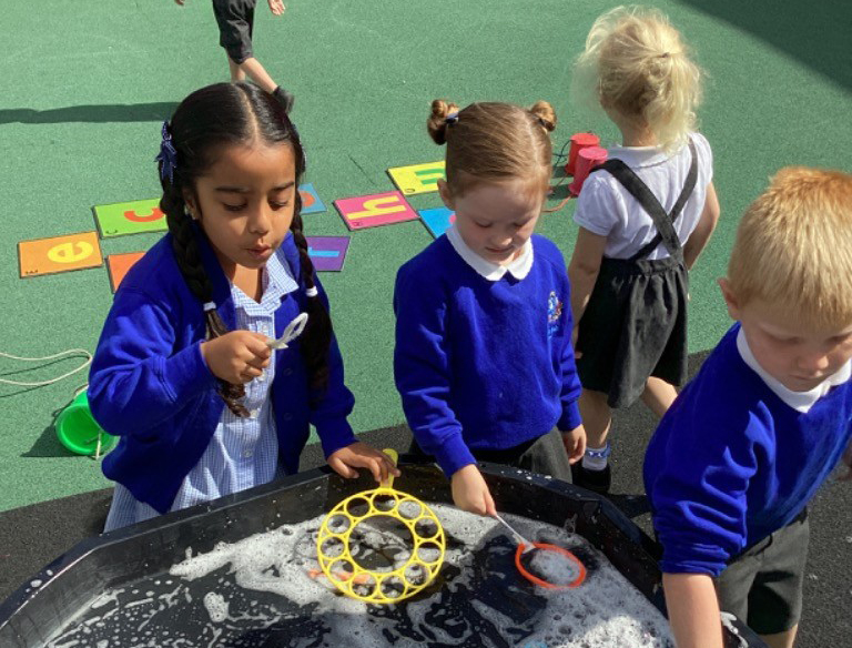 Several children are gathered around a large black tub filled with soapy water and bubbles. Two girls, both wearing blue tops, are actively playing with bubble wands in the water. Another child's arm is visible on the right, also engaging with the water.