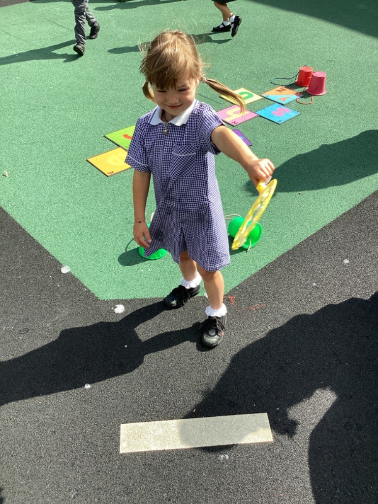 A young girl wearing a blue and white checkered dress is outside on a playground, holding a green bubble wand and appearing to be about to blow bubbles or having just done so. Colorful mats are visible on the green ground behind her.