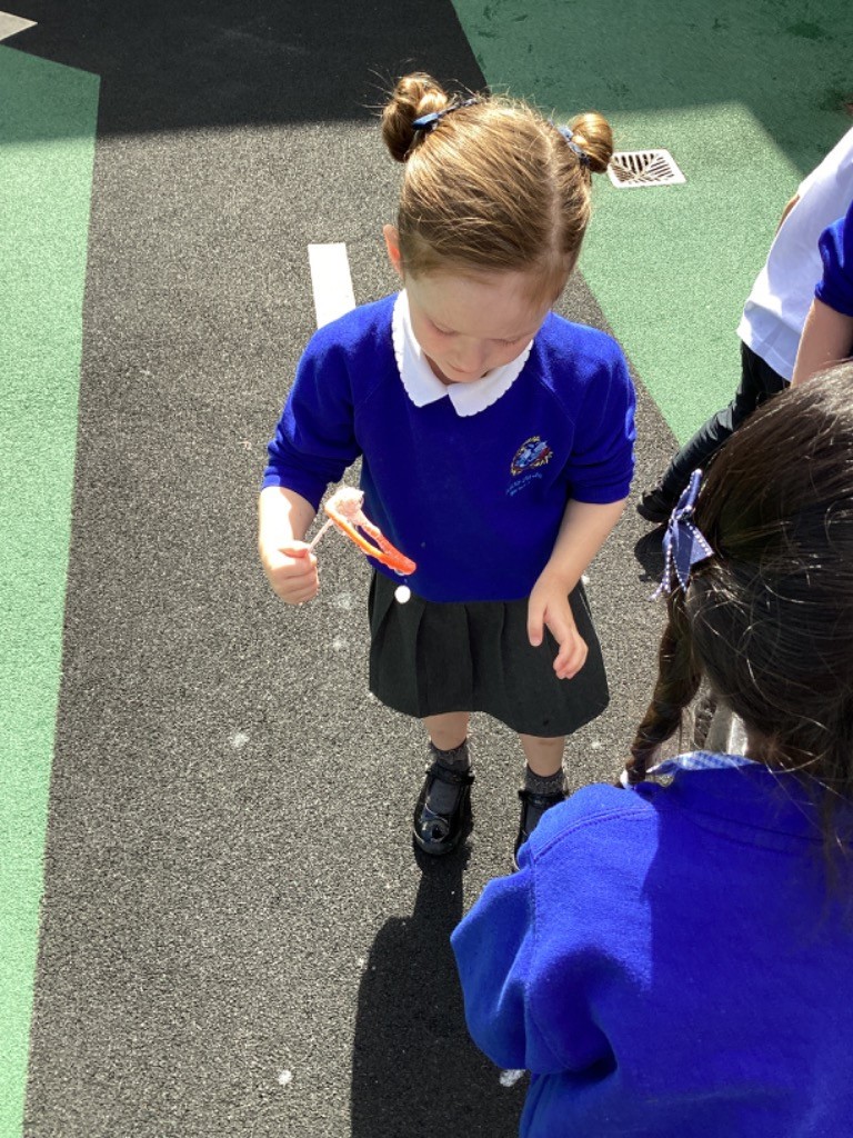 A young girl with her hair in two buns, wearing a blue long-sleeved top and a grey skirt, is playing with a small bubble wand, blowing bubbles. Another child's back is visible in the foreground. They are on a playground surface.