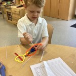 A young boy, wearing a white polo shirt, is seated at a table and appears to be concentrating on a craft activity involving pipe cleaners or similar flexible materials. He is manipulating colorful shapes with his hands.