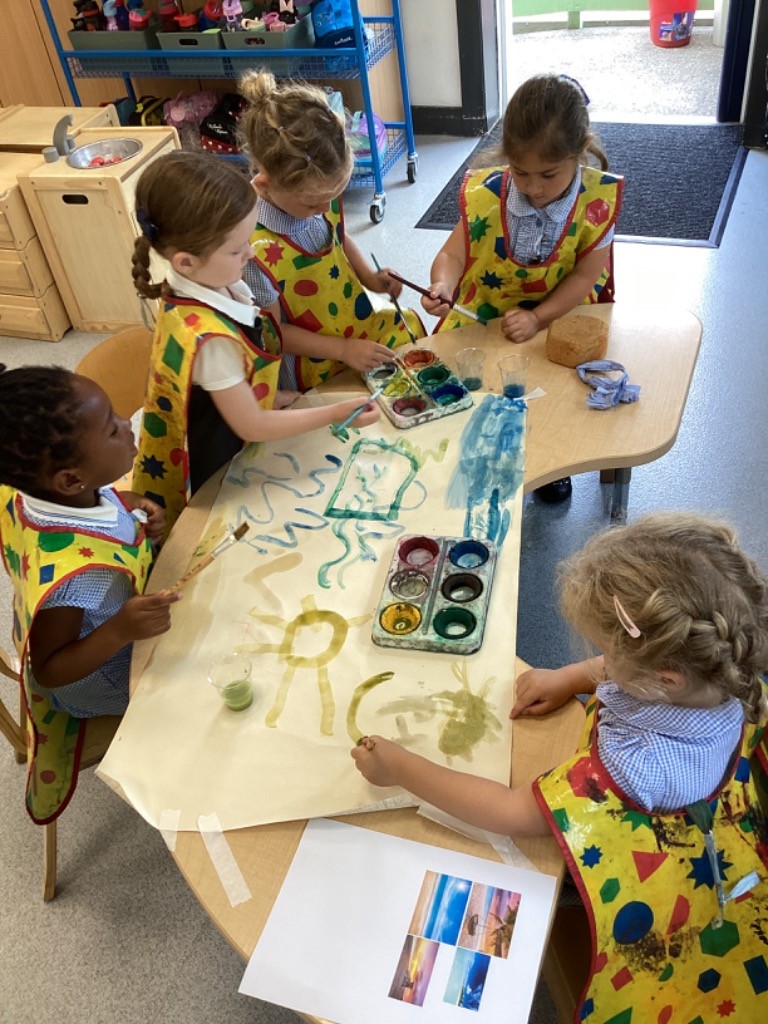 a group of children, all wearing yellow smocks over their school uniforms (blue and white checkered shirts), collaborating on a large sheet of paper with watercolors. They are using paintbrushes and palettes. There are also smaller pieces of paper with images on the table. In the background of LB3, another child is holding up a drawing. These images suggest a creative art session where children are expressing themselves through painting.