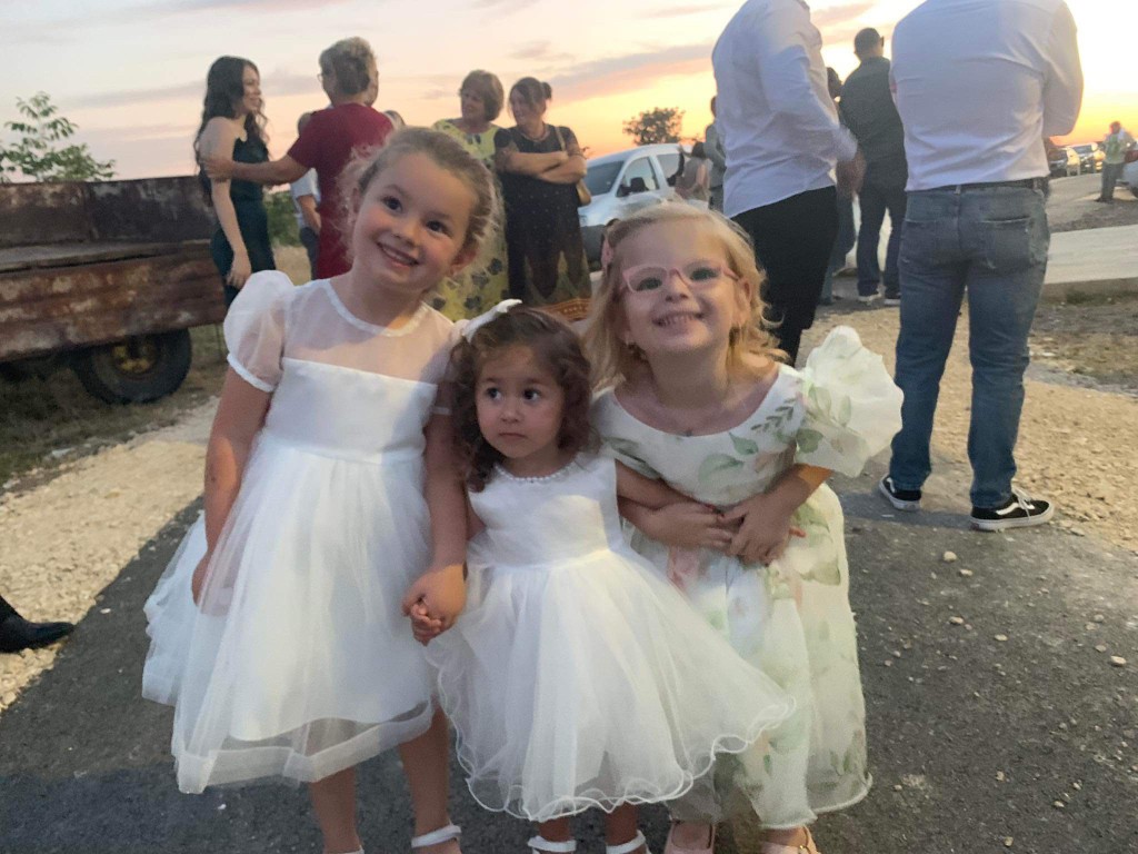 Three young children in white dresses are standing close together and holding hands outdoors. There are adults and a vehicle in the background, suggesting some kind of gathering