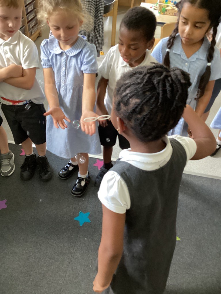 A group of children are gathered around a girl who is blowing bubbles with a bubble wand. Some bubbles are visible in the air and near the children's hands. They are all looking at the bubbles.