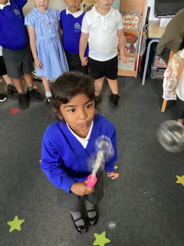 A young girl in a blue top and grey trousers is standing and blowing bubbles with a pink bubble wand. Other children are standing in the background. The floor has yellow star decals.