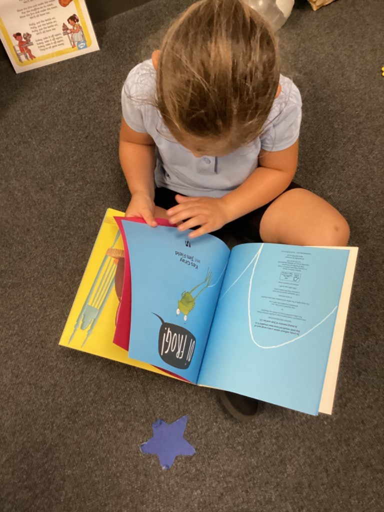 A young child is sitting on a grey carpet, looking at an open copy of the "Oi Frog!" book. The book is open to a page with a blue background and text, along with an illustration of the frog from the cover. A blue star cutout is on the floor next to the book.