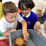 Two young boys, one in a white polo shirt and the other in a blue top, are leaning over a grey tub of water. The boy in the blue top is reaching into the water, possibly touching a brown, hexagonal object. They both seem engaged in the activity.