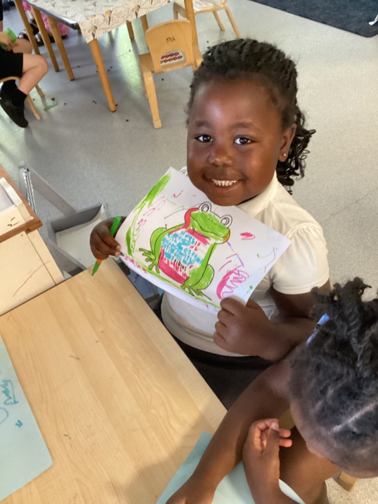 A smiling young girl with dark, braided hair is holding up a colorful drawing of a frog, looking directly at the camera. She's wearing a white polo shirt and is seated at a table, with another child partially visible in the foreground. This appears to be in a classroom setting.