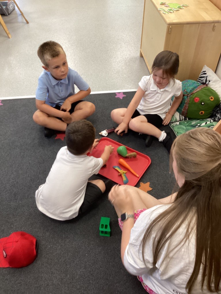 Several children and an adult are sitting on the floor in a classroom. Three children are facing each other, and one child is reaching for items on a red tray that contains toy vegetables and other objects. An adult, mostly visible from the back, is sitting opposite the children. There's a red baseball cap on the floor nearby.