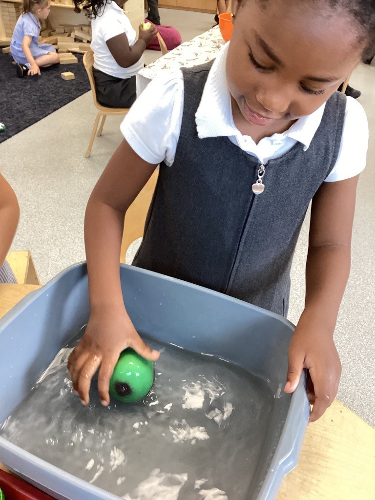 A young girl in a grey pinafore dress and white top is leaning over a grey tub filled with water and is submerging a green, spherical object in it with her hand. She appears to be focused on the activity.