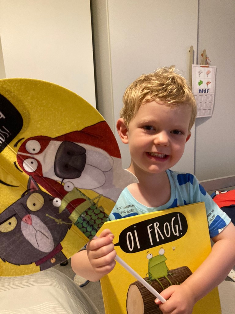A young boy with blond hair is holding the children's book "Oi Frog!" and a large yellow umbrella or parasol with cartoon characters on it, smiling broadly at the camera. He's wearing a striped t-shirt and appears to be indoors.