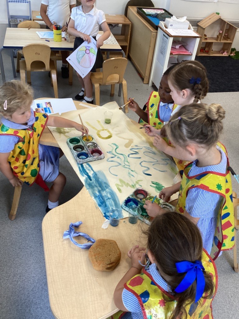 a group of children, all wearing yellow smocks over their school uniforms (blue and white checkered shirts), collaborating on a large sheet of paper with watercolors. They are using paintbrushes and palettes. There are also smaller pieces of paper with images on the table. In the background of LB3, another child is holding up a drawing. These images suggest a creative art session where children are expressing themselves through painting.
