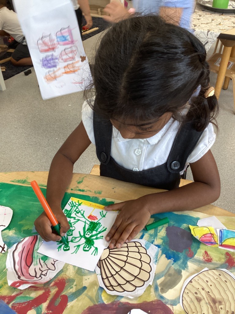 This picture shows a child with long dark hair, wearing a white shirt and dark overalls, intently drawing with an orange marker on a small piece of paper. There are also cut-out shapes, including a seashell, on the table, indicating a creative or craft activity.