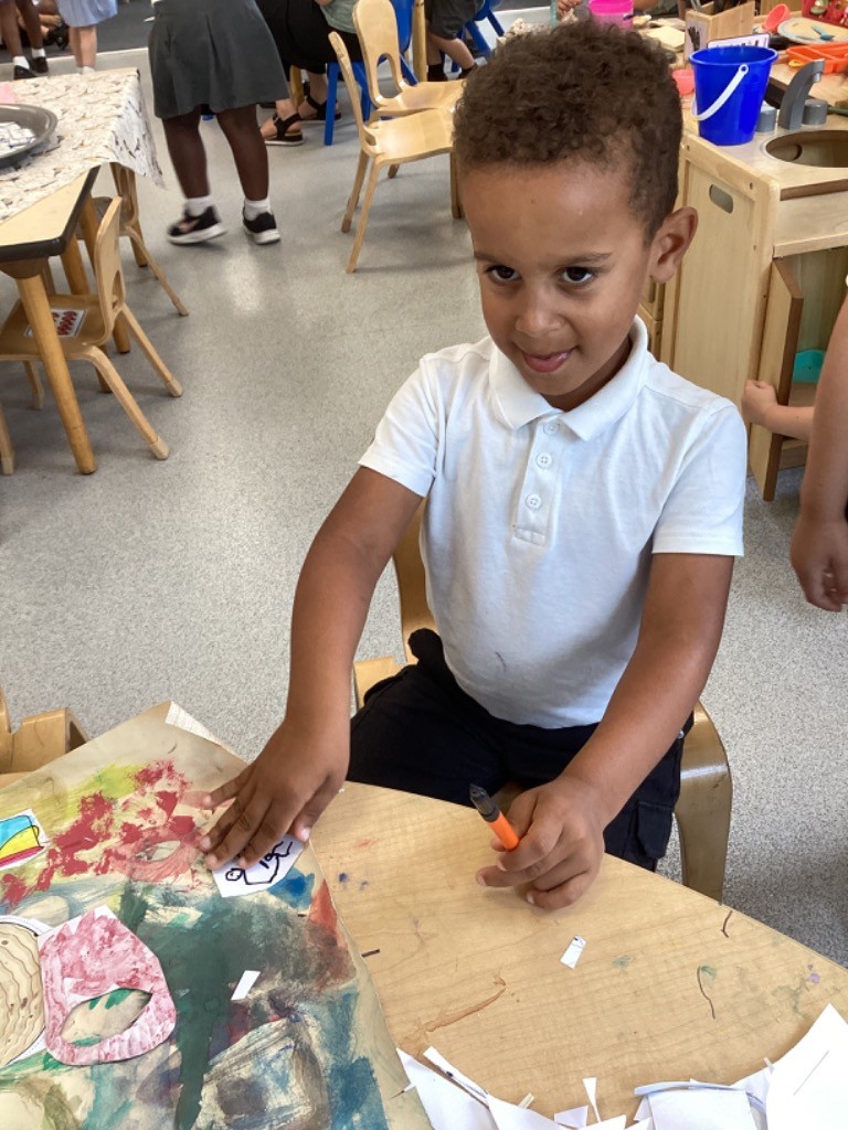 This image features a young boy with curly hair, wearing a white polo shirt, sitting at a table with drawings and crafts. He is holding an orange marker and looking towards the camera with a slight smile, suggesting he is engaged in a creative activity.