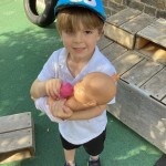 A child wearing a white polo shirt, black shorts, and a blue cap is smiling at the camera while holding a doll and a pink bottle. This appears to be outdoors.
