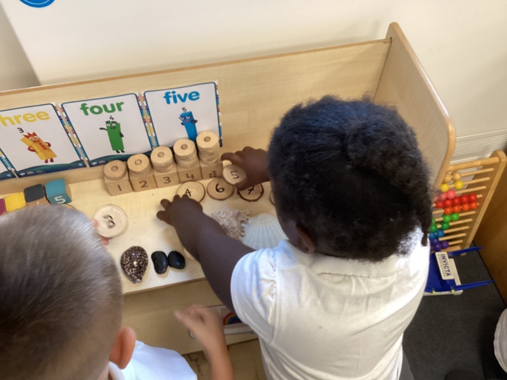 children at a learning station focused on numbers and counting. They are interacting with wooden blocks stacked with numbers (1 to 5), wooden discs with numbers, seashells, and polished stones. In the background of LB7, another child is playing with building blocks. These activities suggest early numeracy learning, possibly involving counting, number recognition, and one-to-one correspondence using natural materials. Posters with numbers and characters (like "three," "four," "five") are visible on the wall.