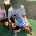 A child wearing a white polo shirt and a green baseball cap is sitting outdoors, holding a doll wrapped in a blue blanket.