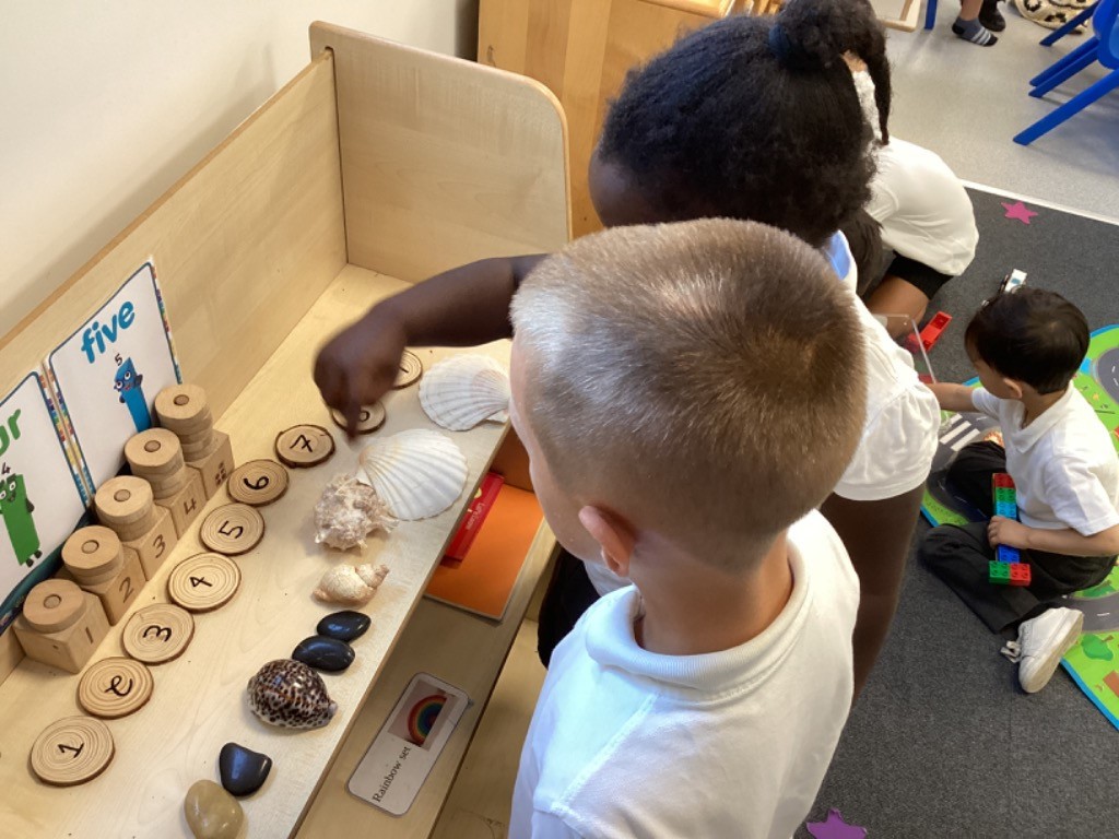 children at a learning station focused on numbers and counting. They are interacting with wooden blocks stacked with numbers (1 to 5), wooden discs with numbers, seashells, and polished stones. In the background of LB7, another child is playing with building blocks. These activities suggest early numeracy learning, possibly involving counting, number recognition, and one-to-one correspondence using natural materials. Posters with numbers and characters (like "three," "four," "five") are visible on the wall.