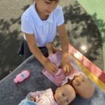 A child in a white polo shirt is standing over two dolls on a grey mat, appearing to be playing with them. A pink bottle is also on the mat.
