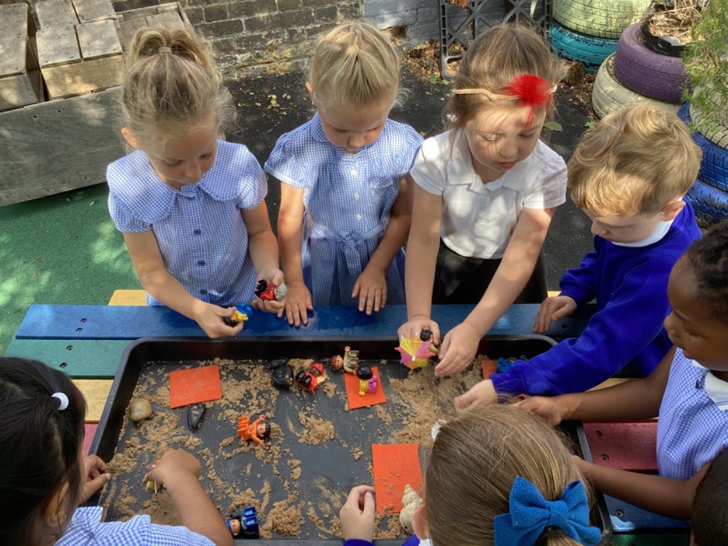 several children, some in blue long-sleeved tops and some in blue and white checkered dresses, gathered around a large black tray filled with sand and various small objects like small figures, orange squares, and tiny umbrellas. They are actively engaged in manipulating the sand and objects, suggesting imaginative or sensory play with a focus on fine motor skills. LB8 shows more children involved, and one child has a red feather in their hair.