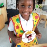 A smiling child wearing a colorful art smock is holding up a paper plate with what appears to be a craft or painting on it.