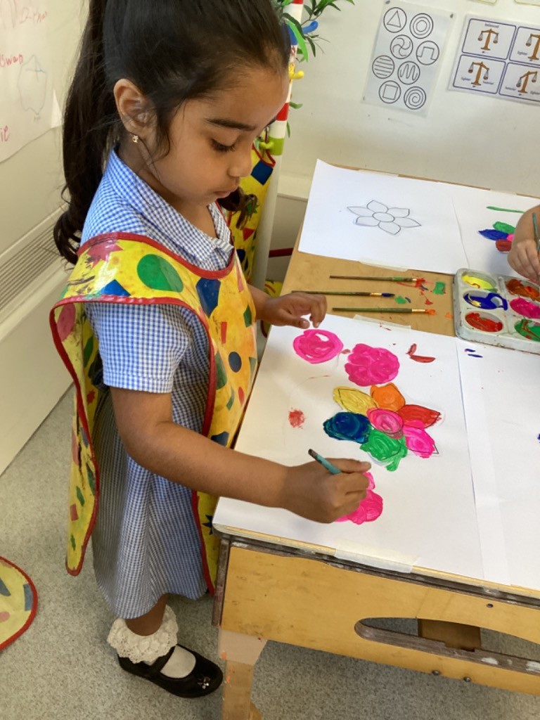 A young girl, wearing a checkered school uniform and a yellow art smock, is standing at a table and painting. She is focused on her artwork, which depicts colorful shapes, possibly flowers, on a white sheet of paper. There are paintbrushes and a paint palette on the table.