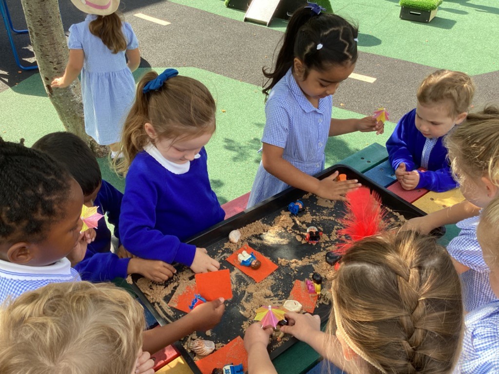 several children, some in blue long-sleeved tops and some in blue and white checkered dresses, gathered around a large black tray filled with sand and various small objects like small figures, orange squares, and tiny umbrellas. They are actively engaged in manipulating the sand and objects, suggesting imaginative or sensory play with a focus on fine motor skills. LB8 shows more children involved, and one child has a red feather in their hair.