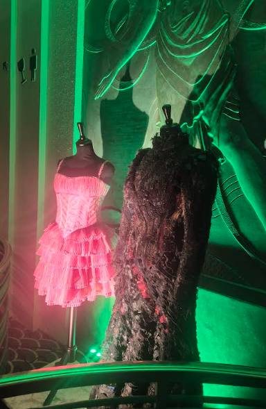 Two mannequins are on display, each wearing an elaborate costume. One costume is a pink, ruffled dress, and the other is a dark, textured, possibly organic-looking garment. The lighting in the scene is green. This looks like a costume display, possibly for a theatrical production.
