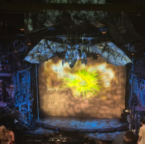 This image shows a stage set, likely for a theatrical performance. A large map titled "The Land of Oz" is projected or painted onto the backdrop, and a large, dragon-like prop hangs above it. The set design features gears and industrial elements. This strongly suggests a scene from a production of "Wicked" or a similar show set in the world of Oz.