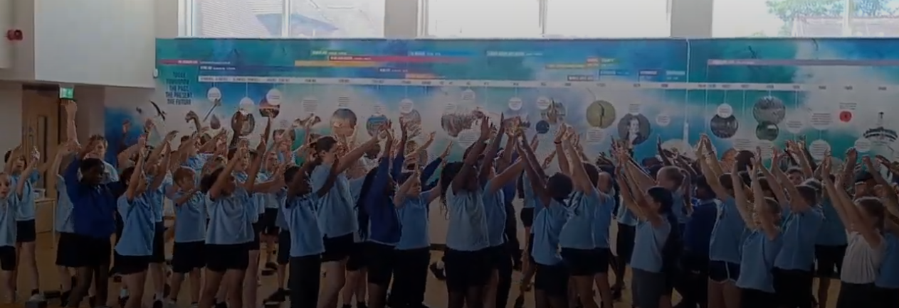 A large group of children, likely students, are gathered indoors, possibly in a school hall or gymnasium. They are all raising their arms above their heads. There's a large mural or display on the wall behind them that appears to be a timeline.