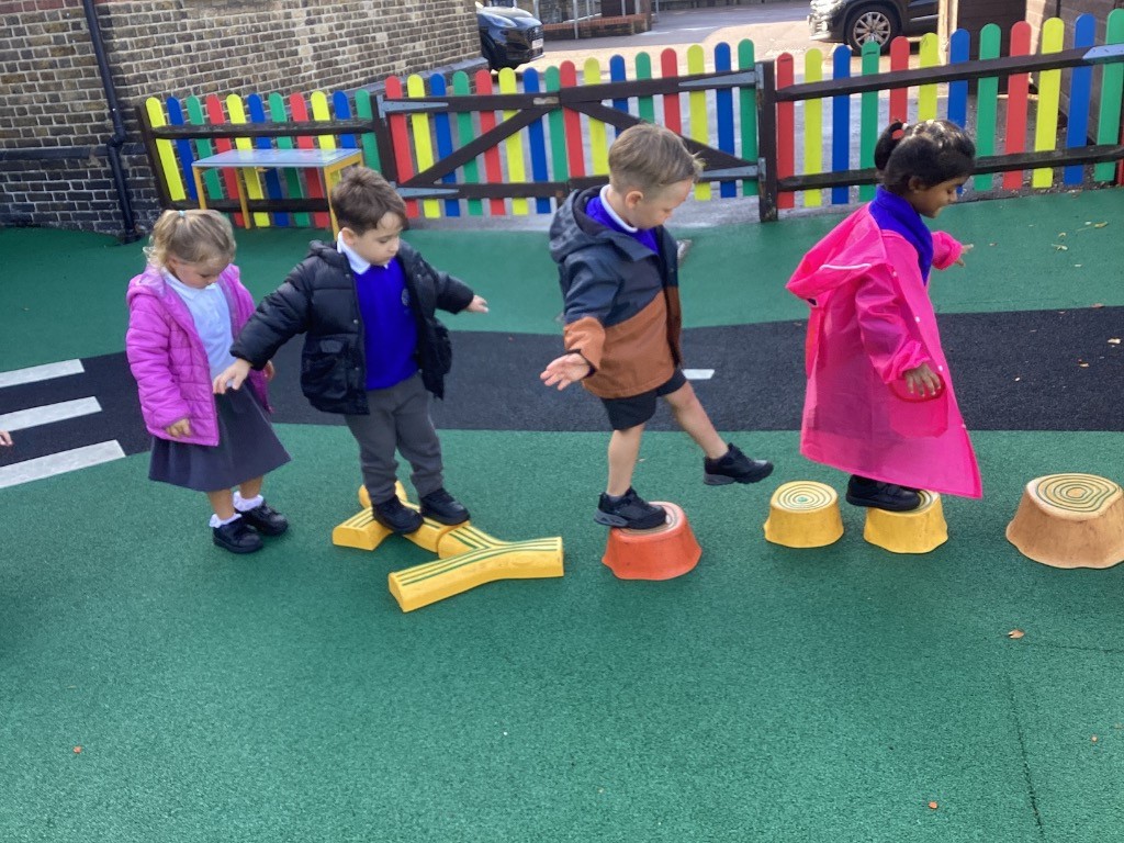A group of children is playing on an outdoor playground. They are walking and balancing on a series of brightly colored stepping stones and a wooden beam.