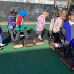 Several children are outside, walking along a line of wooden blocks, seemingly balancing as they move from one block to the next.