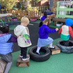 Several children are playing outside, walking and balancing on a variety of objects, including wooden blocks and large tires.