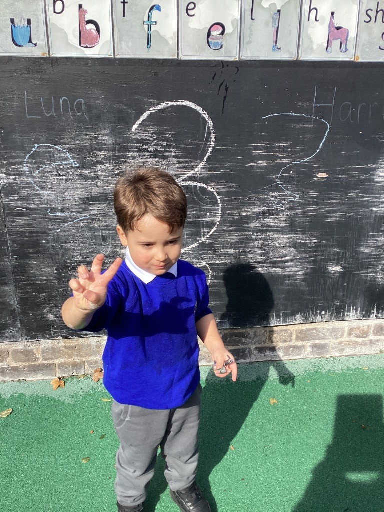 A child is standing in front of a large black chalkboard, holding up two fingers and looking down at their hand. The numbers "3" and "4" are written on the board, along with some names.