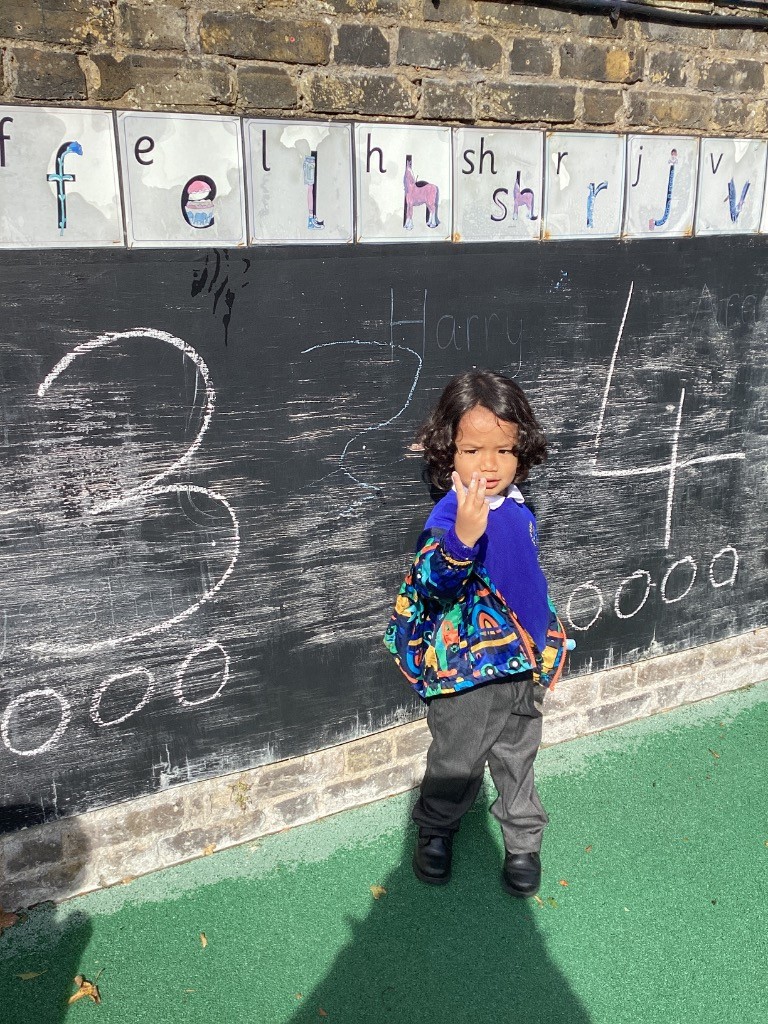 A child is standing in front of a large black chalkboard, holding up one hand and looking directly at the camera. Numbers and names are written on the board behind them.