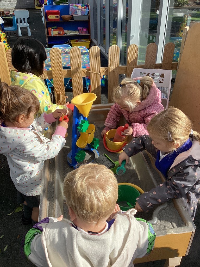 Several children are gathered around a sensory table, actively engaged in play. They are using various toys, including a water wheel, funnels, and buckets, to interact with the contents of the table.