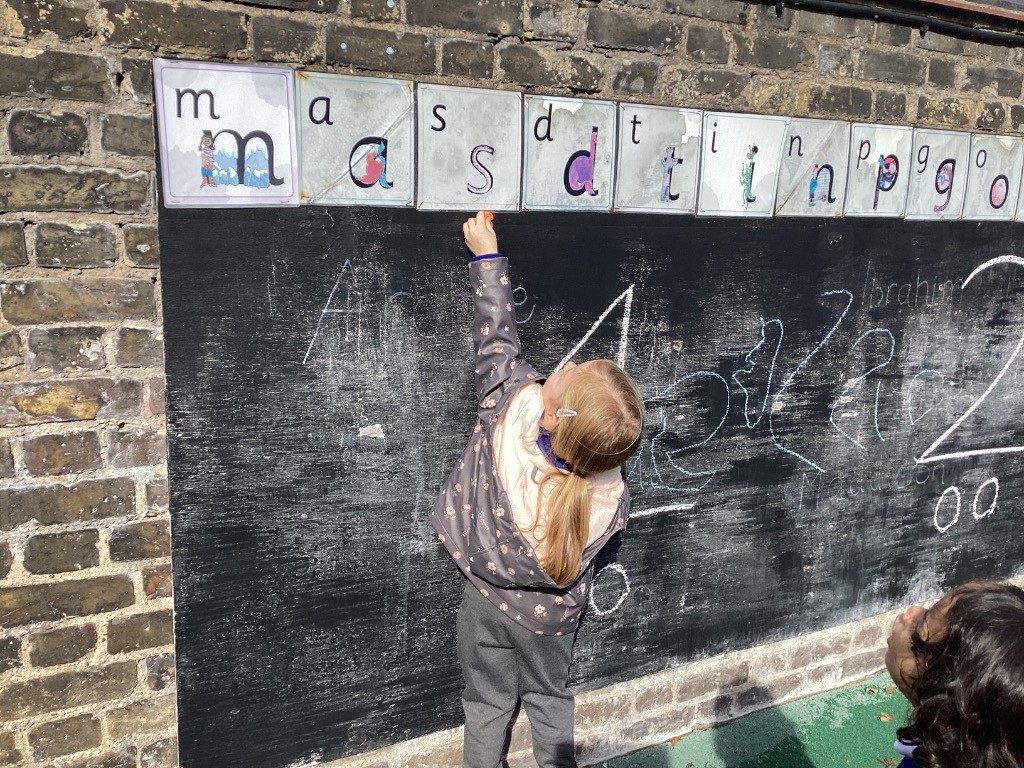 A child is reaching up and pointing at one of the alphabet cards hanging above the chalkboard while holding a piece of chalk.
