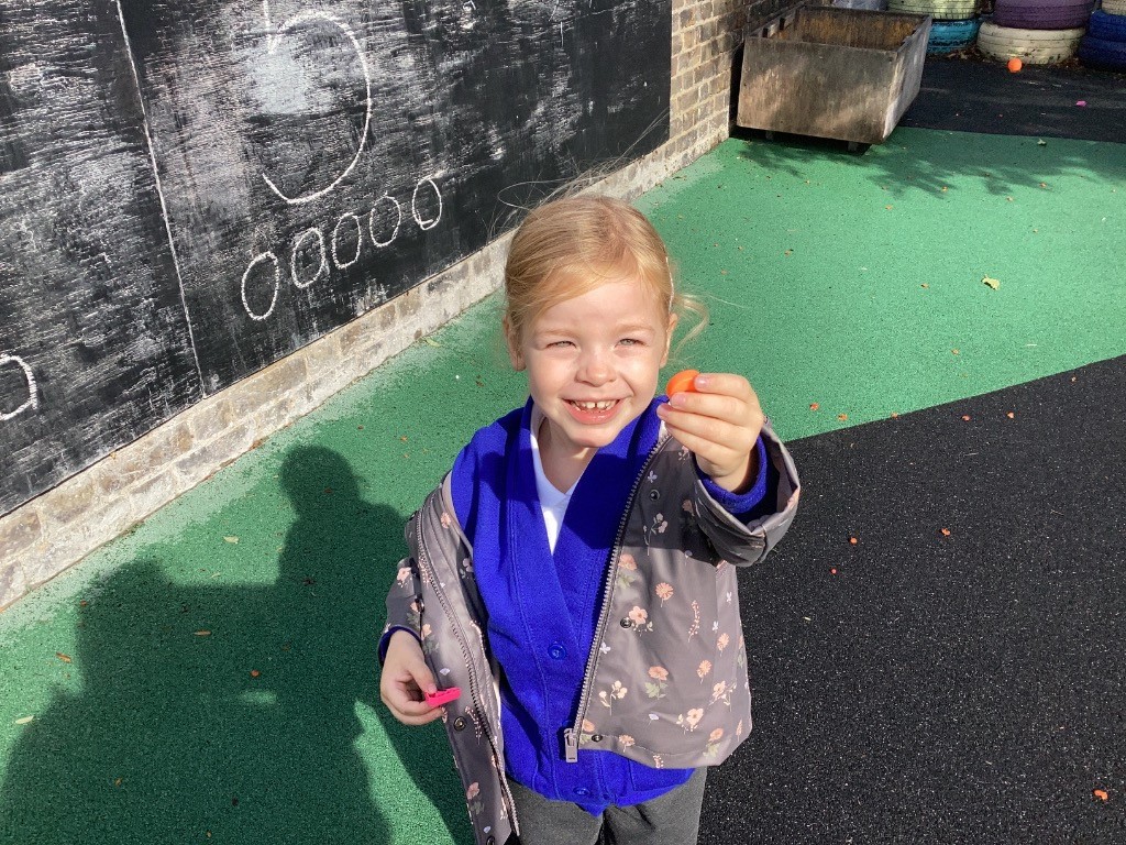 A child is smiling at the camera, holding a small, orange object in one hand and a pink object in the other. They are standing in front of the same chalkboard seen in the other images.