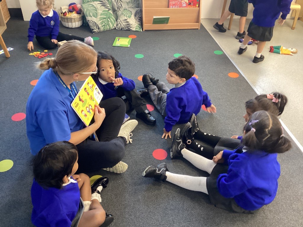 An adult is sitting on the floor with a group of children, reading a book aloud to them. They are all gathered together in what looks like a classroom setting.