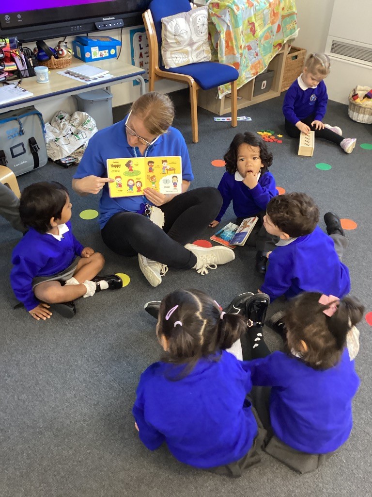 An adult is sitting on the floor with a group of children, reading a book aloud to them. They are all gathered together in what looks like a classroom setting.