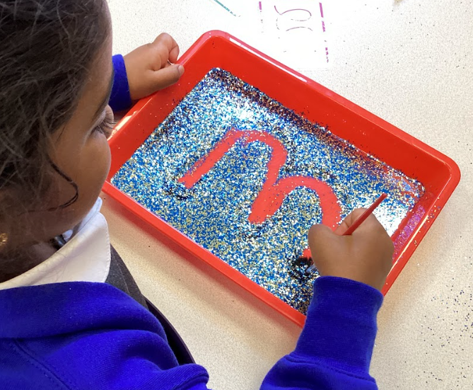 This image shows a young child using a brush to trace a large letter "m" in a red tray filled with blue and gold glitter. This is a sensory activity designed to help children learn to form letters in a creative, tactile way. The child is using a brush to carefully draw the letter in the glitter.