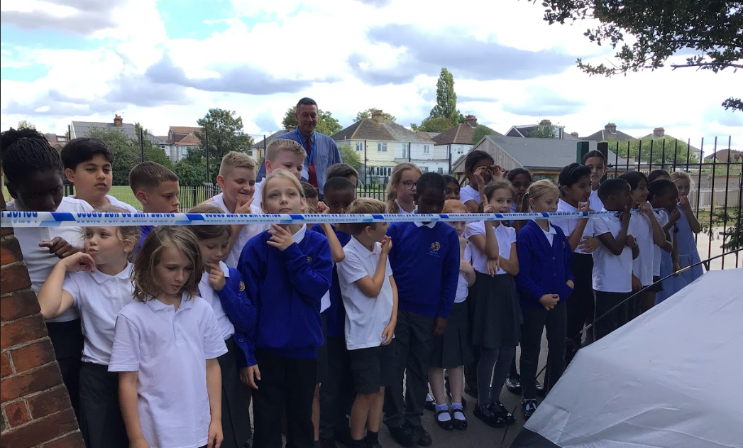 a group of students is gathered outside, observing what appears to be a mock crime scene. The area is cordoned off with blue and white "Police Line - Do Not Cross" tape.