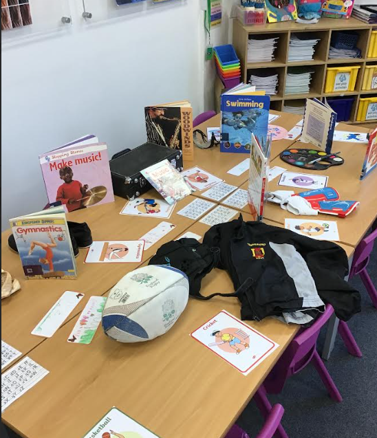 a table is set up with various resources for learning about different sports, including books on gymnastics and swimming, a rugby ball, and cards with images of cricket and basketball.