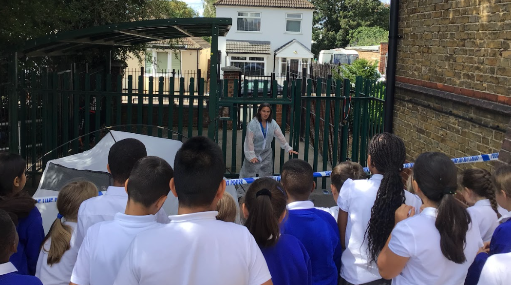 an adult, possibly a teacher or presenter, is dressed in what looks like a forensic suit, pointing and explaining something to the students. This suggests they are learning about forensics or a similar topic through an engaging, hands-on experience.