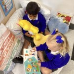 two children are relaxing together in a cozy corner with pillows and stuffed toys, looking at a book.