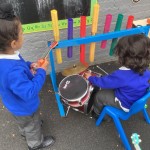 two children are engaging in a musical activity, one is playing a drum and the other is playing a xylophone-like instrument made of colorful tubes.