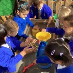a group of children are participating in a group activity around a tray, using various tools and containers to play with a substance like sand or water.